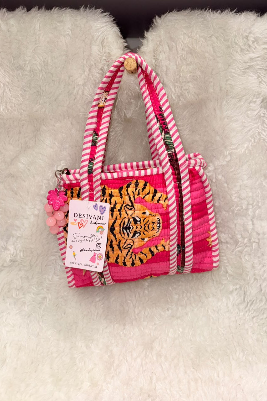 A small pink and white striped fabric handbag with a tiger face patch on the front, hanging on a white faux fur surface. The bag has pink-and-white striped handles, a pink and white striped zipper pull, and a hanging paper tag that reads 'DESVANI'.