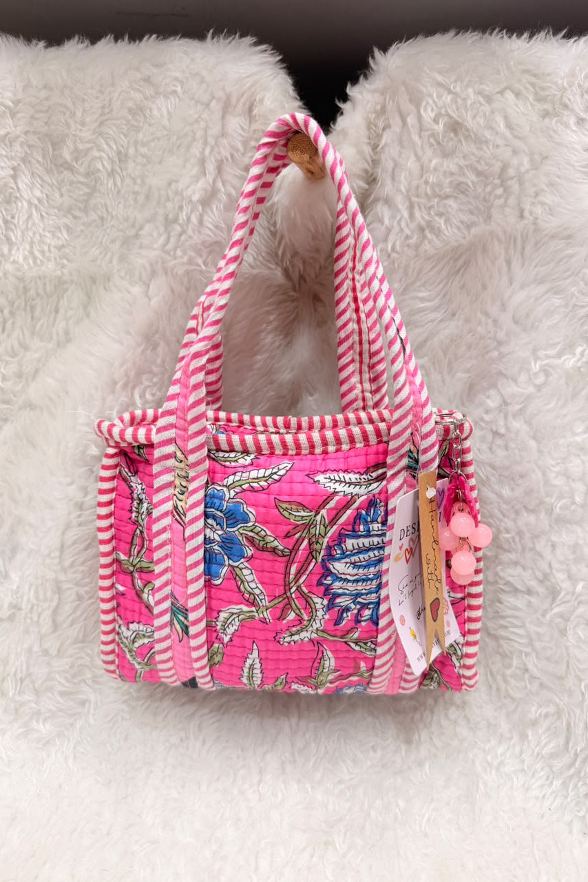 A small pink tote bag with a floral print and pink-and-white striped handles, resting on a white faux fur surface.