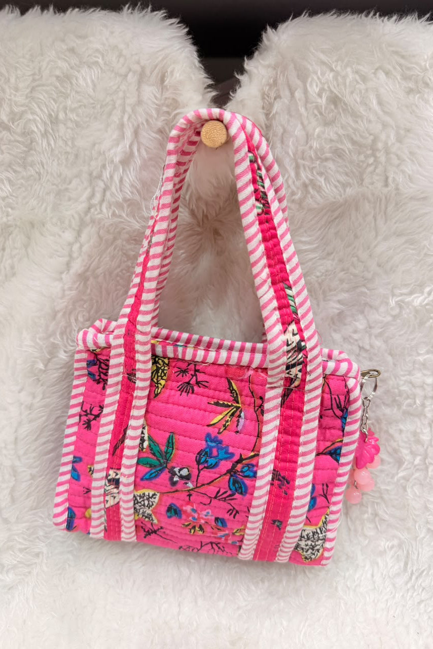 A small quilted tote bag in bright pink with white striped handles and trim, decorated with a colorful floral and bird print, resting on a white faux fur surface.