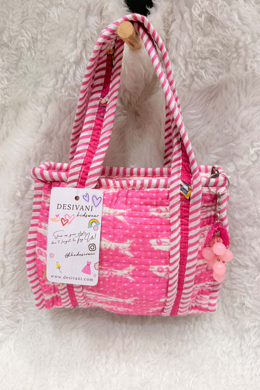 A pink and white striped fabric tote bag with a quilted front pocket, hanging on a white faux fur surface. The bag has pink pom-pom charms on the side and a paper brand tag that reads 'DESIVANI handmade'.