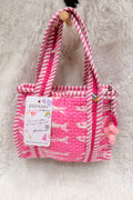 A pink and white striped fabric tote bag with a quilted front pocket, hanging on a white faux fur surface. The bag has pink pom-pom charms on the side and a paper brand tag that reads 'DESIVANI handmade'.