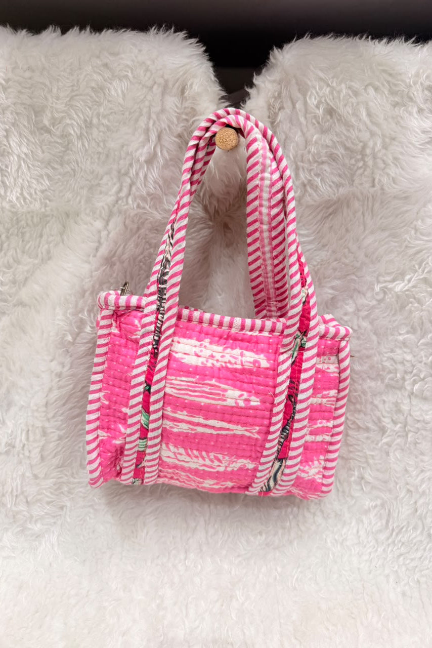 A small woven tote bag in bright pink and white striped fabric, hanging from a wooden hook against a white faux fur background.